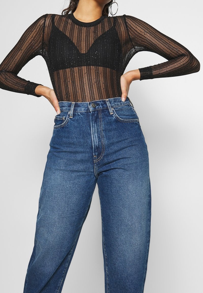 Bodysuit noir transparent avec des rayures verticales et des accents brillants, associé à un jean en denim bleu taille haute avec une coupe classique et cinq poches.