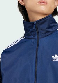 Primo piano di una persona che indossa una giacca sportiva Adidas blu con il colletto chiuso e strisce bianche sulla spalla.
