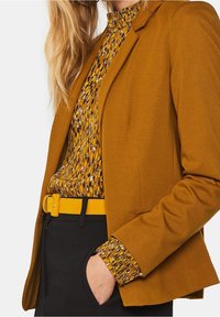 Hořčičná sako s přizpůsobeným designem, hladkým materiálem a dlouhými rukávy. Nosí se přes vzorovanou hořčičnou a černou blůzu, doplněnou žlutým páskem.