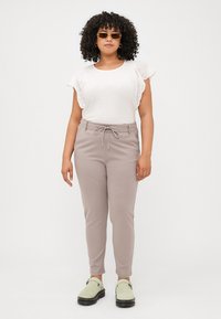 Eine Frau mit lockigem Haar steht selbstbewusst in einem neutralen Hintergrund und trägt ein weißes Oberteil, beigen Hosen und grüne Clogs.