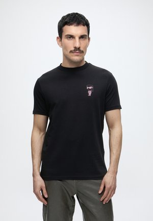 Homme avec moustache portant un t-shirt noir avec un petit dessin animé rose sur la poitrine et un pantalon gris, debout devant un fond uni.