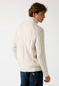 Maglione color crema con colletto alto, caratterizzato da motivi a treccia testurizzati e a coste. Indossato con jeans blu scuro, che evidenziano una vestibilità rilassata.