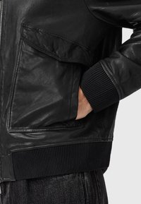 Sort læderbomberjakke med lynlås, sidelomme og ribstrikkede sorte manchetter. Glat tekstur med subtil glans.