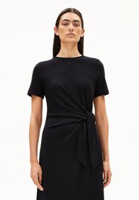 Schwarzes Kleid aus weichem Material. Es hat kurze Ärmel, einen runden Ausschnitt und ein geknotetes Detail in der Taille, das eine fließende Silhouette erzeugt.
