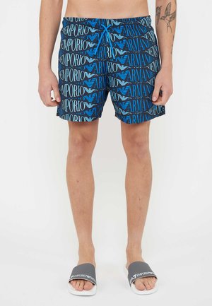 Man draagt blauwe en zwarte Emporio Armani zwembroek met logopatroon en donkergrijze en witte Emporio Armani slippers.