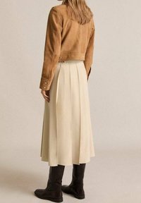 Bruine suède cropped jas, beige geplooide midi-rok en zwarte kniehoge laarzen gezien van achteren. Soepele texturen en nauwsluitende vormen.