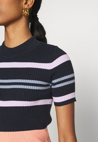 Lyhythihainen ribbiknitti-toppi, jossa on vaakasuorat raidat tummansinisessä, vaaleansinisessä, vaaleanpunaisessa ja persikassa, sekä puolikorkea kaula-aukko. Näkyvissä kultaiset rengaskorvakorut.