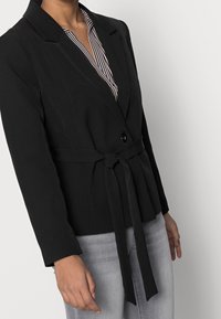 Zwarte blazer met ceintuur, lange mouwen, enkele knoopsluiting en een V-hals, gedragen over een gestreept shirt. Glad stof en een getailleerde pasvorm.