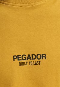 Gul t-skjorte i bomull med ribbestruktur, med svart trykt tekst: "PEGADOR" og "BUILT TO LAST" i fet skrift.