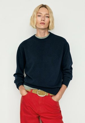 Person med kort blondt hår iført en marineblå sweater, røde bukser og et lysebrunt bælte, står med den ene hånd i lommen foran en ensfarvet baggrund.