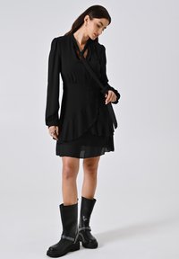 Schwarzes langärmliges Kleid mit einem gerafften Saum, Bindegürtel und durchsichtigen Schichten. Kombiniert mit kniehohen schwarzen Stiefeln mit Nietenakzenten.