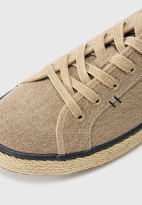 Beige lerret sneaker med rund tå, forsterkede snorfester og brune snører. Juteomslag på sålen, svart gummitrim langs den nederste kanten.