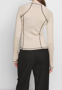 Beige lange mouw top van rekbaar, glad materiaal met contrasterende zwarte stiksels. Voorzien van een ronde halslijn en een getailleerd ontwerp.