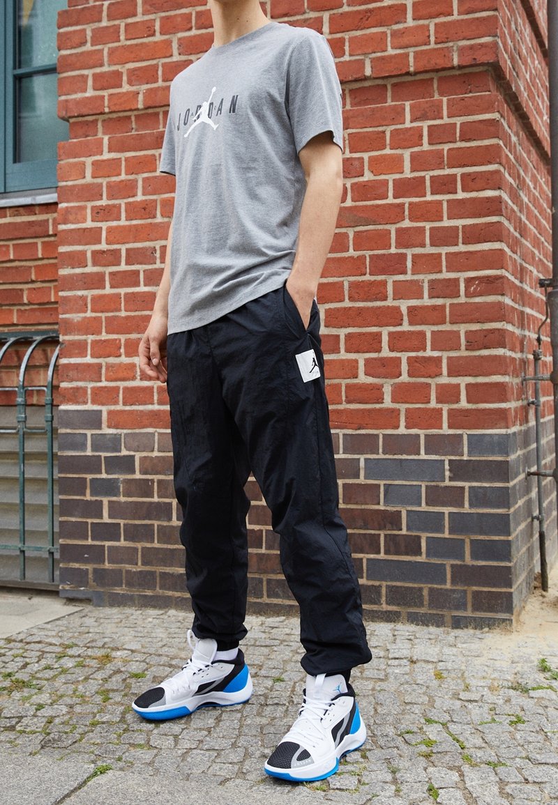 Grå bomullströja med "Jordan"-logotyp, svarta nylon joggers med sidofickans logotyp, och svarta/blåa sportskor med vita accenter.