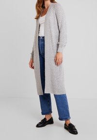 Šedý pletený cardigan s Chevron texturou, otevřený vpředu a s žebrovanými manžetami, kombinovaný s modrými džínami s vysokým pasem a černými mokasínami.