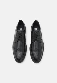 Chaussures brogues en cuir noir avec des perforations décoratives, un bout arrondi et une semelle épaisse. Les lacets sont noirs avec une finition lisse.