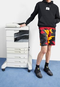 Persona con felpa nera con cappuccio e pantaloncini colorati a zigzag che tocca una grande stampante per ufficio bianca su un tappeto blu con sfondo di parete bianca.