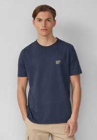 Jonge man met lichtbruin haar, gekleed in een marineblauw t-shirt met de tekst "BEAT THE HEAT" en beige shorts, staande tegen een grijze muur.