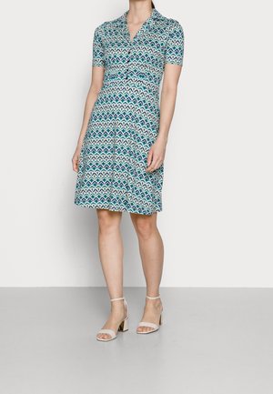 Woman wearing knee-length, short-sleeve dress with blue and green geometric pattern, standing, paired with white block-heeled sandals.