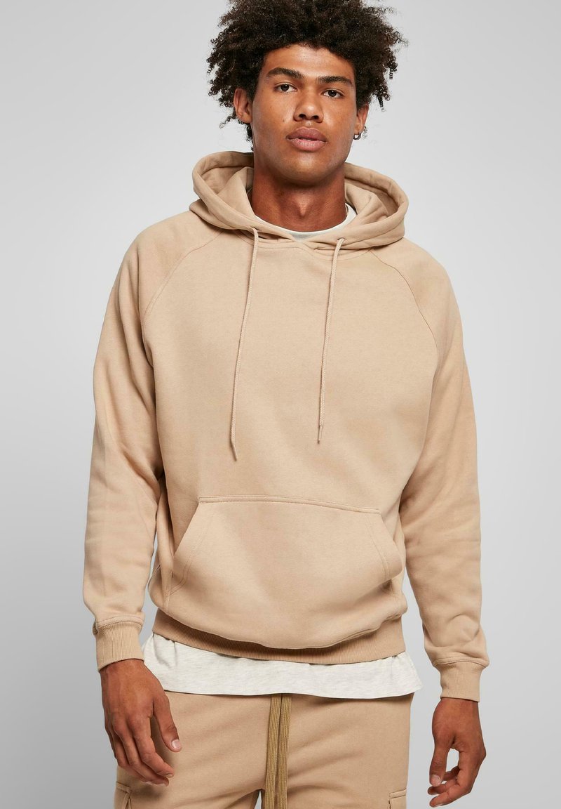 Felpa beige realizzata in tessuto morbido, con tasca a marsupio, cappuccio con coulisse e polsini a coste. Indossata sopra una maglietta grigia chiara.