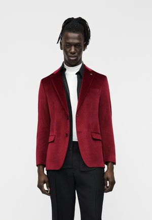 Muž s dredmi oblečený v červenom zamatovom saku cez čierne tričko a biely rolák, v kombinácii s čiernymi nohavicami, stojaci pred jednoduchým pozadím.