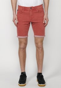 Shorts de mezclilla rojos con dobleces en los puños, cinco bolsillos y un corte regular. Combinados con zapatillas negras, que muestran un look casual.