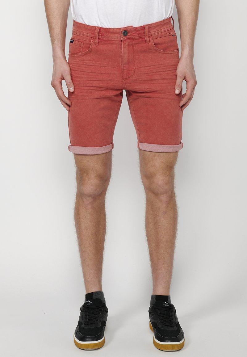 Shorts de mezclilla rojos con dobleces en los puños, cinco bolsillos y un corte regular. Combinados con zapatillas negras, que muestran un look casual.