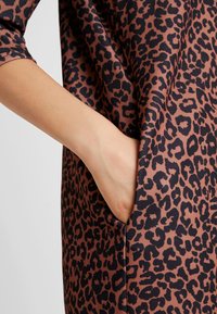 Käsi resting in side pocket of brown dress with black leopard print, showing three-quarter sleeve ja smooth fabric texture.