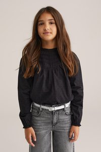 Svart långärmad blus med draperad hals, texturerad tyg och subtil prickmönster. Kombineras med grå jeans och ett ljust bälte.