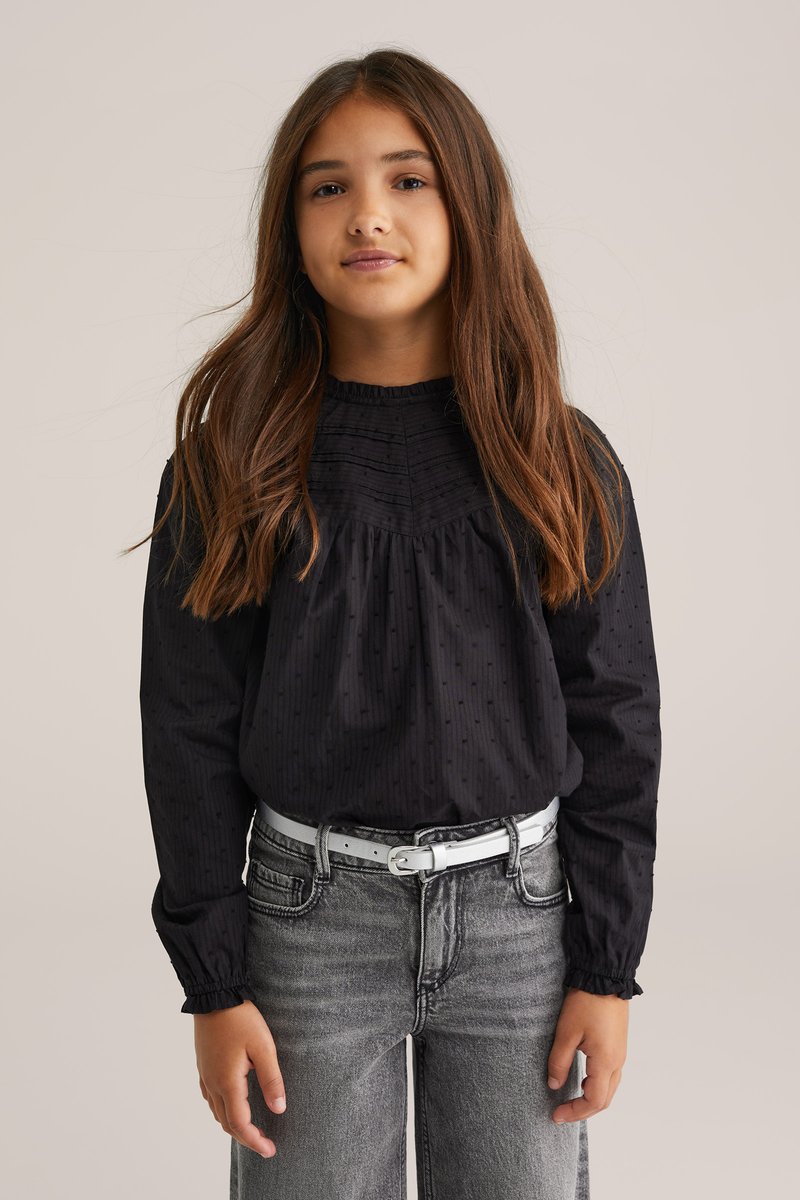 Svart långärmad blus med draperad hals, texturerad tyg och subtil prickmönster. Kombineras med grå jeans och ett ljust bälte.