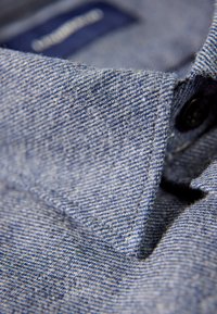 Blå skjorta med krage i texturerat tyg, med ett diagonalt vävmönster och svart knappstängning. Söm synlig längs kragen.