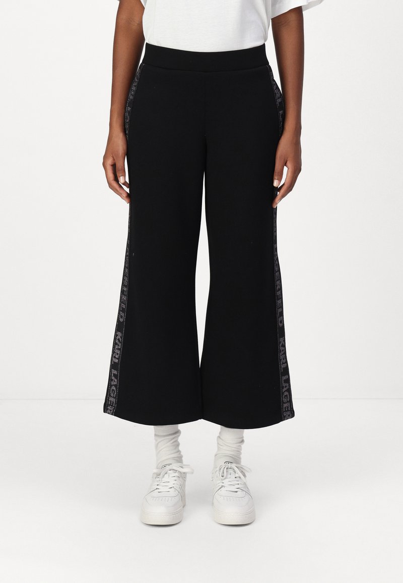 Fekete, bő szárú melegítőnadrág derékkal, ezüst logóval a szélén, fehér tornacipővel és fehér zoknival párosítva.