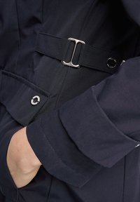 Veste bleu marine avec ceinture à boucle argentée, poche latérale avec garniture circulaire, et tissu texturé présentant un motif subtil en chevron.