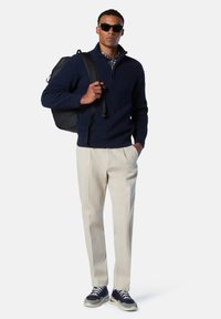 Pullover in maglia a coste navy con mezza zip, pantaloni plissettati crema e sneakers scure con dettagli rossi. Aggiunti uno zaino nero e occhiali da sole.
