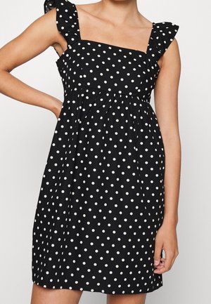 Woman wearing a sleeveless black dress with white polka dots and ruffle shoulder straps, hand resting on hip.