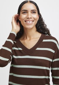 Pull en maille à rayures brunes avec un décolleté en V. Comprend des rayures horizontales vert d'eau claire et un tissu en tricot texturé. Modèle souriant, touchant ses cheveux.