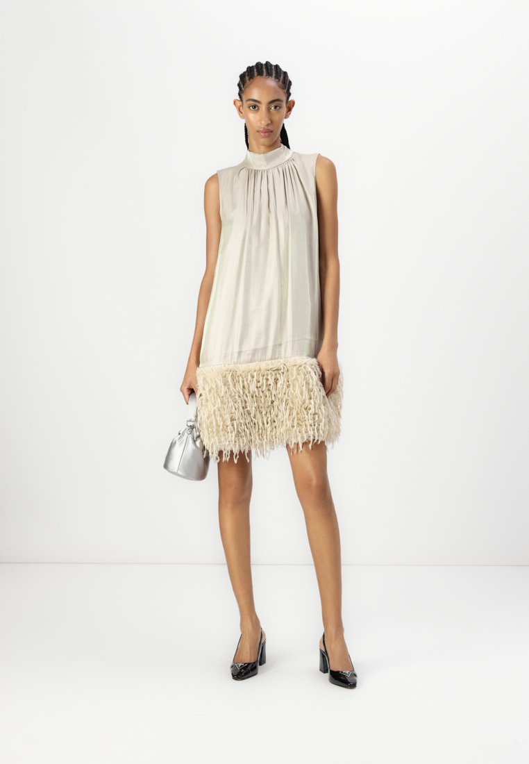 Robe sans manches beige avec un col froncé et une texture à franges au niveau de l'ourlet. Assortie à un sac à main argenté et des talons noirs.