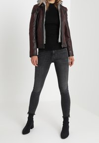 Braune Lederjacke mit grauem Innenfutter, schwarzer gerippter Pullover, schwarze ausgefranste Jeans und schwarze Stiefeletten, die vor einem neutralen Hintergrund stehen.