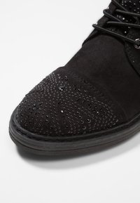 Botín de ante negro hasta el tobillo con punta redondeada, que presenta un exterior texturizado adornado con pequeños detalles brillantes en negro y una suela de goma.