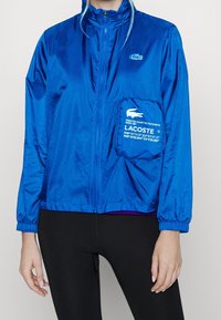 Blå nylonjacka med dragkedja fram, elastiska ärmslut och en framficka med vit Lacoste-logotyp och koordinater. Slät, glänsande yta.