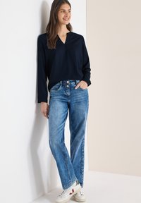 Haut bleu marine à manches longues avec un col en V, associé à un jean bleu clair et des baskets blanches avec des accents rouges. Le mannequin s'appuie contre un mur.