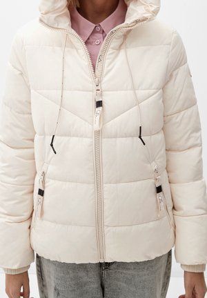 Vrouw draagt een crèmekleurige gewatteerde donsjas met capuchon, ritssluiting aan de voorkant, zijzakken met rits en een roze overhemd met kraag eronder.