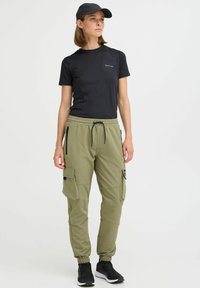 Black fitted t-shirt, olive green cargo pants with elastic cuffs and zip pockets, paired with black sneakers and a matching cap.
