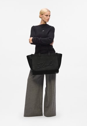 Zwarte leren tote bag met een gestructureerd ontwerp en een geperst "Karl" logo, voorzien van twee handvatten. Grijze wijde broek en zwarte trui.