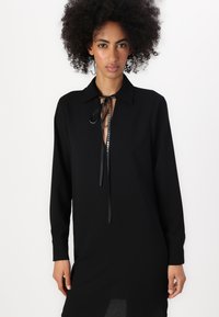 Vestito nero con scollatura a colletto e maniche lunghe. Presenta una chiusura a cravatta con paillettes decorative e una vestibilità leggermente più ampia.