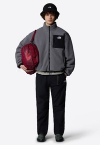 Giacca in pile grigia con chiusura a zip, tasca nera. Cappello a secchiello nero, borsa a tracolla rossa, pantaloni neri, scarpe in camouflage. Design semplice e funzionale.