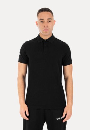 Mand står vendt fremad iført en ensfarvet sort kortærmet poloshirt og sorte bukser med hvid tekst på venstre lår.