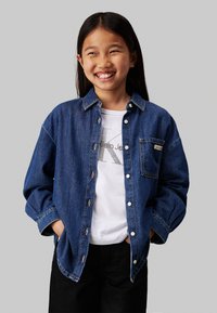 Camicia di jeans, blu scuro, con bottoni frontali, grandi tasche, maniche arrotolate, indossata sopra una maglietta bianca con un logo grigio, abbinata a pantaloni neri.