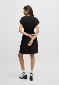 Zwarte jurk met korte mouwen, smock taille, lengte tot de knie, gemaakt van een gestructureerde stof, gecombineerd met zwarte sandalen met hak en witte sokken.