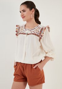 White blouse with colorful embroidered pattern and ruffled sleeves, paired with orange shorts. Soft fabric, button-up design, casual fit.
